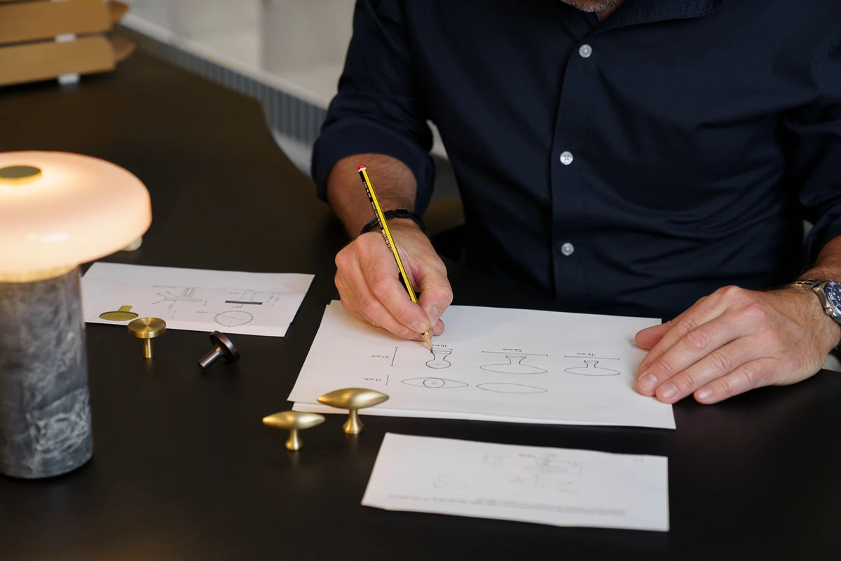 En person tegner møbelknopper ved et skrivebord med produktprøver og en tændt bordlampe.