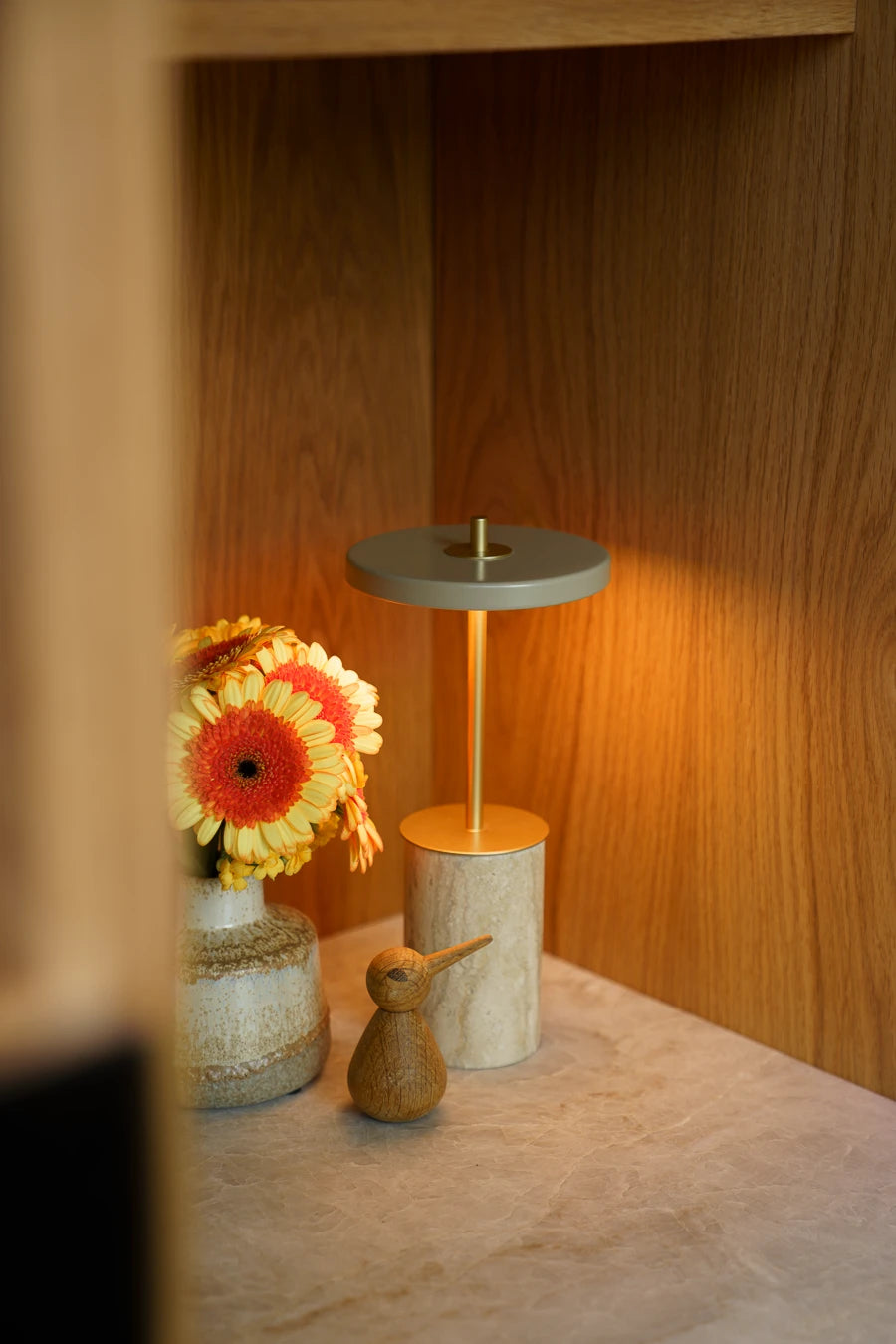 Tændt Asteria Move Mini bordlampe med marmorfod ved en vase med orange blomster i en træniche.