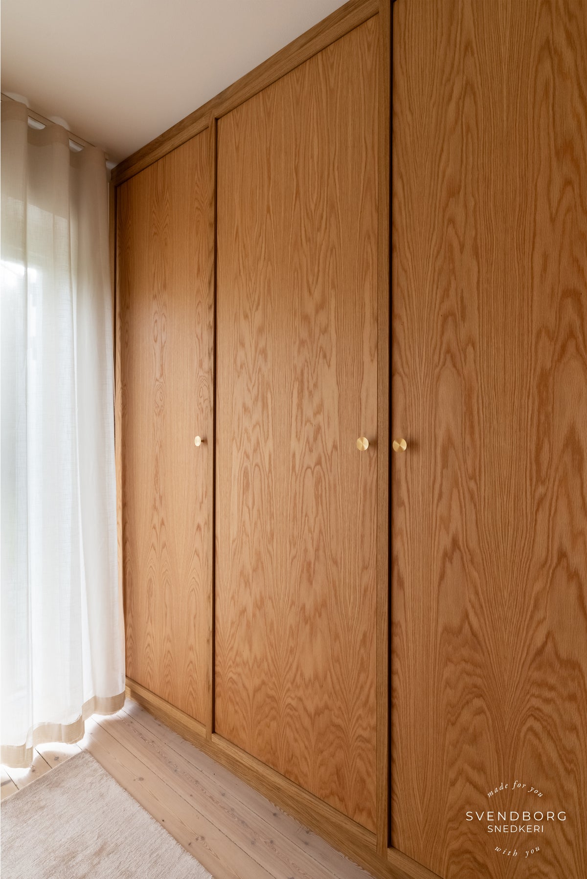 Høj trægarderobe med runde København-knopper i messing ved lyse gardiner.