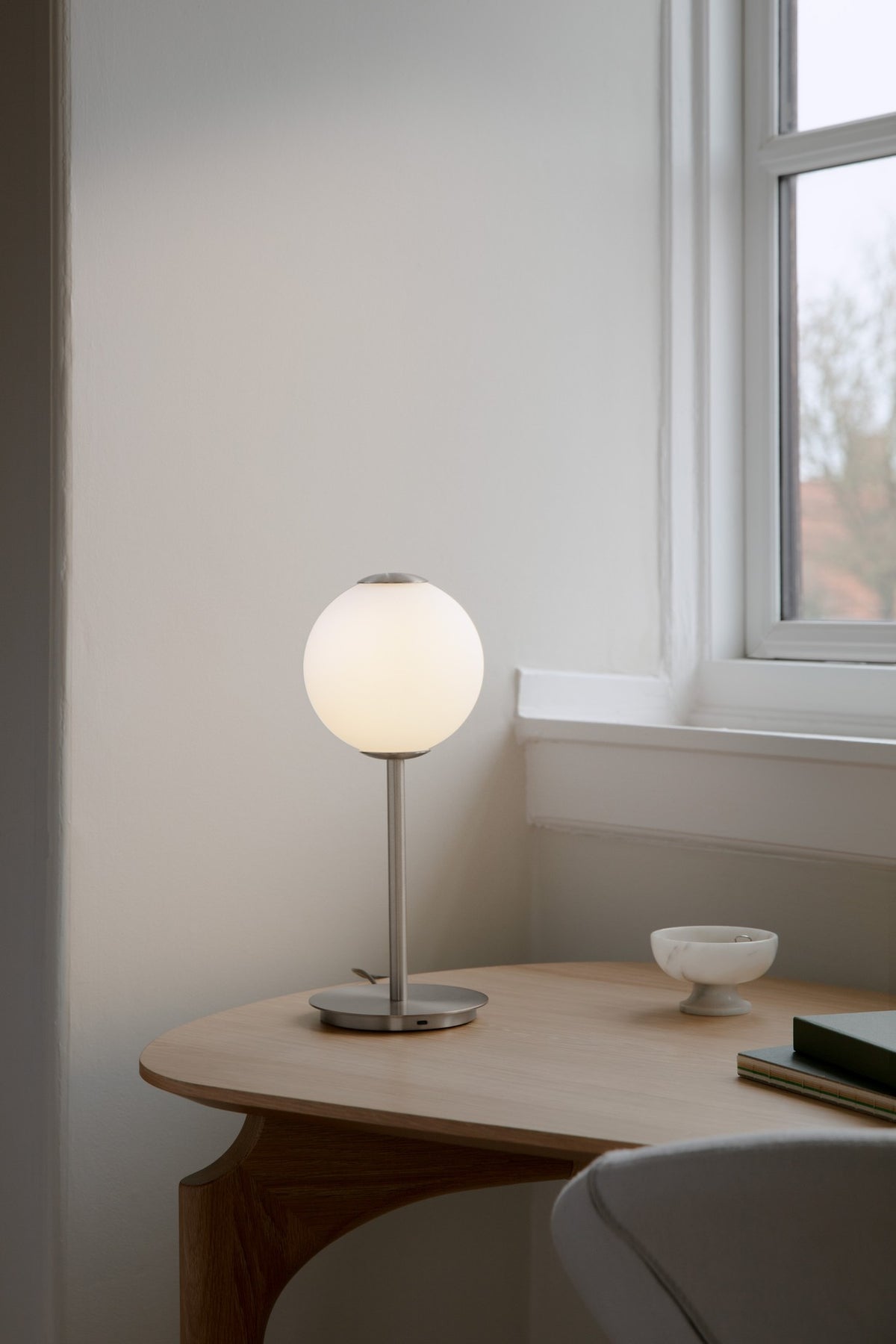 Audrey Table bordlampe i stål med lysende kugleskærm på et træbord ved et vindue.