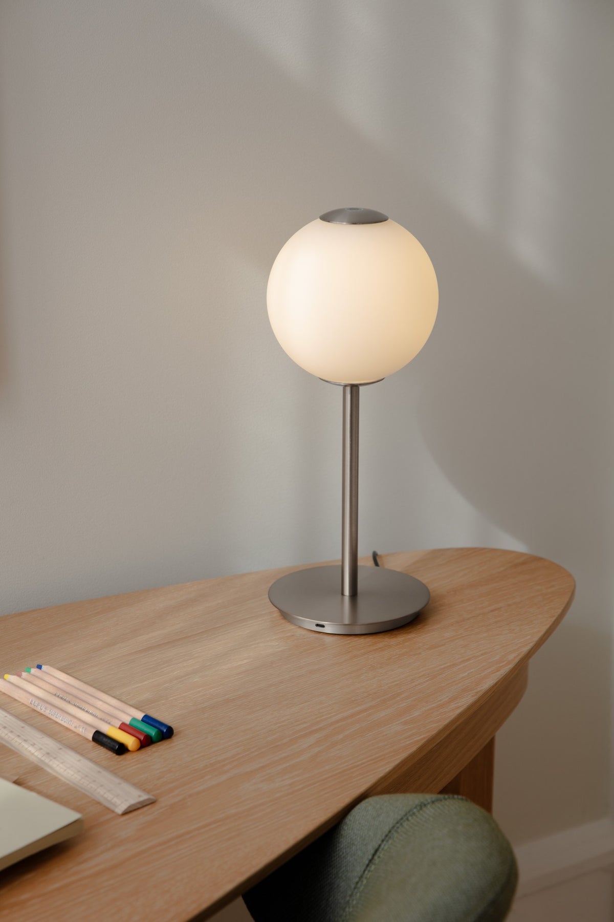 Tændt Audrey Table bordlampe i stål fra UMAGE på et træbord med farveblyanter.